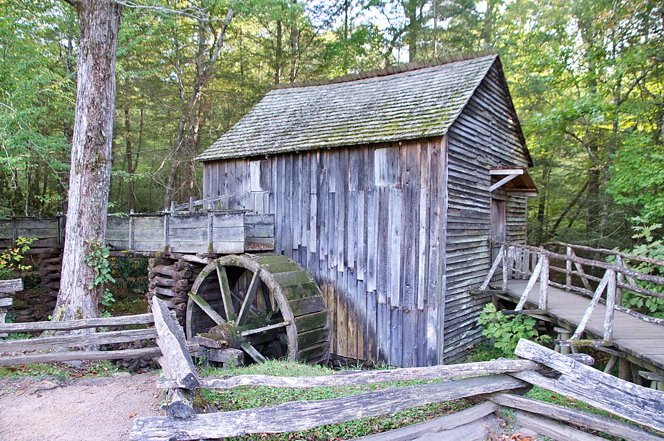 Cable Mill at Cades Cove in Great Smoky Mountains National Park, Tennessee. The historic grist mill features a moss-covered wooden water wheel and weathered board-and-batten siding, framed by a split rail fence and surrounded by dense forest. The mill still operates today and is one of the most visited historic structures in the park