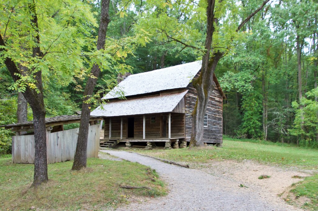 Historic wooden Carter Shields cabin with a shingled roof, set among trees along a gravel path in Cades Cove