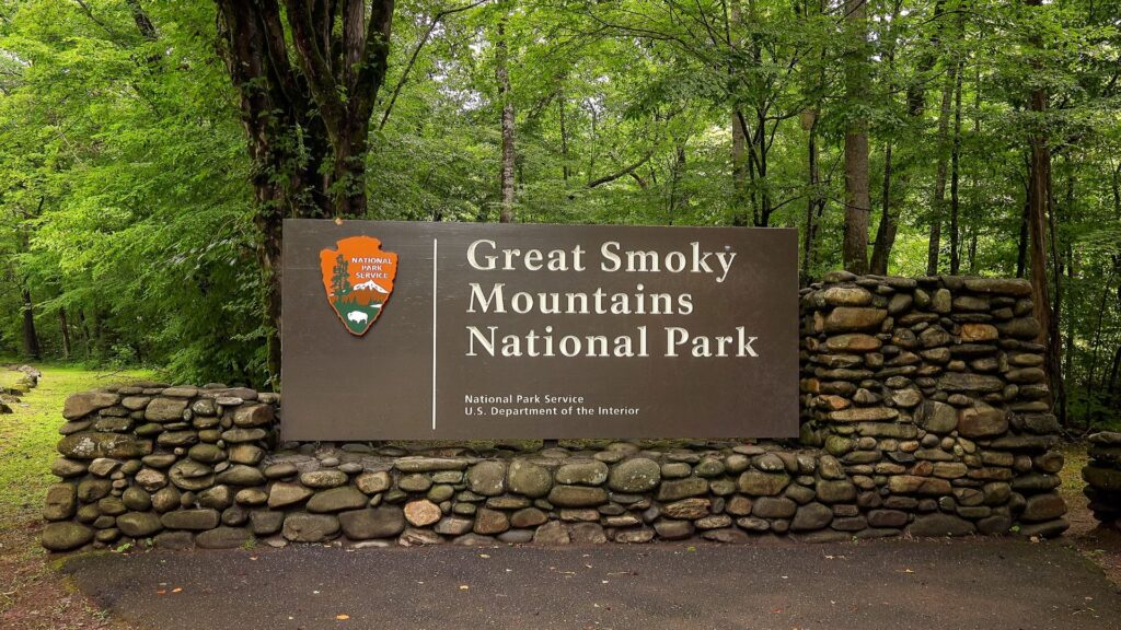The Great Smoky Mountains National Park entrance sign with the National Park Service arrowhead logo, set into a stone wall surrounded by green forest