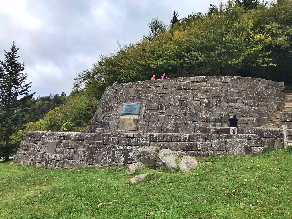 A large two-tiered stone monument at Newfound Gap with visitors standing atop it, surrounded by forest under a cloudy sky