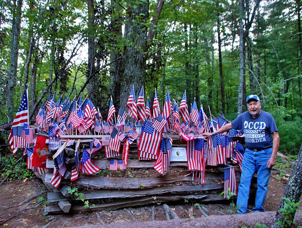 Sam Buchanan stands beside the Pearl Harbor Tree memorial at Cades Cove in Great Smoky Mountains National Park, Tennessee. The fallen tree stump is covered with dozens of small American flags and patriotic tributes left by visitors to honor those who died at Pearl Harbor on December 7, 1941, creating a spontaneous woodland memorial deep in the Smoky Mountains forest
