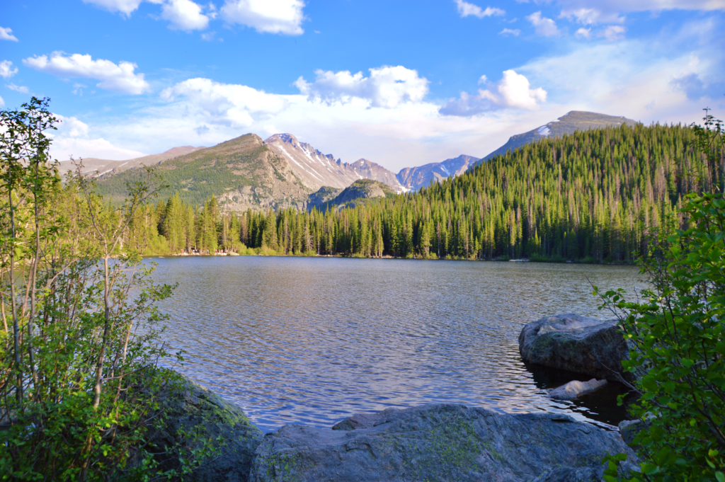 A calm lake surrounded by dense pine forest, boulders in the foreground, and rugged mountain peaks with patches of snow rising against a partly cloudy sky