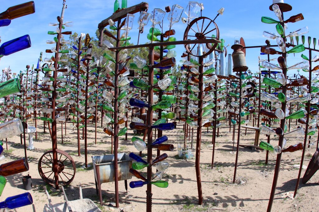 A dense forest of handmade metal trees covered in hundreds of colorful glass bottles, vintage lanterns, and old wheels stands in a sandy desert yard under a clear blue sky
