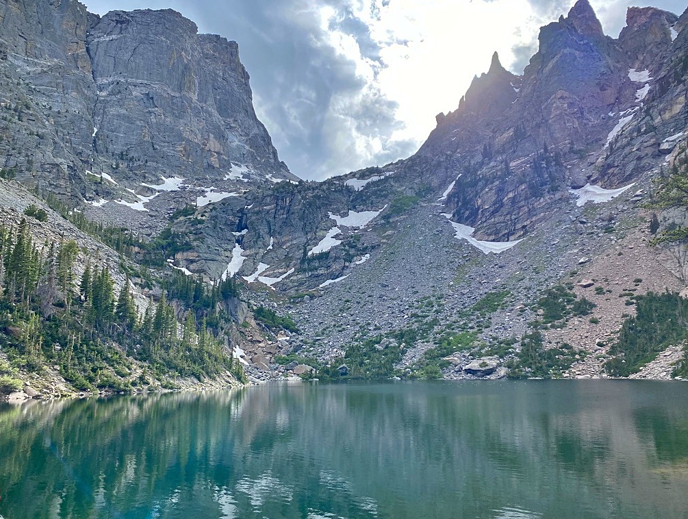 A turquoise alpine lake surrounded by pine trees and rocky shoreline, with the steep, snow-streaked face of Hallett Peak rising against a partly cloudy sky in the background
