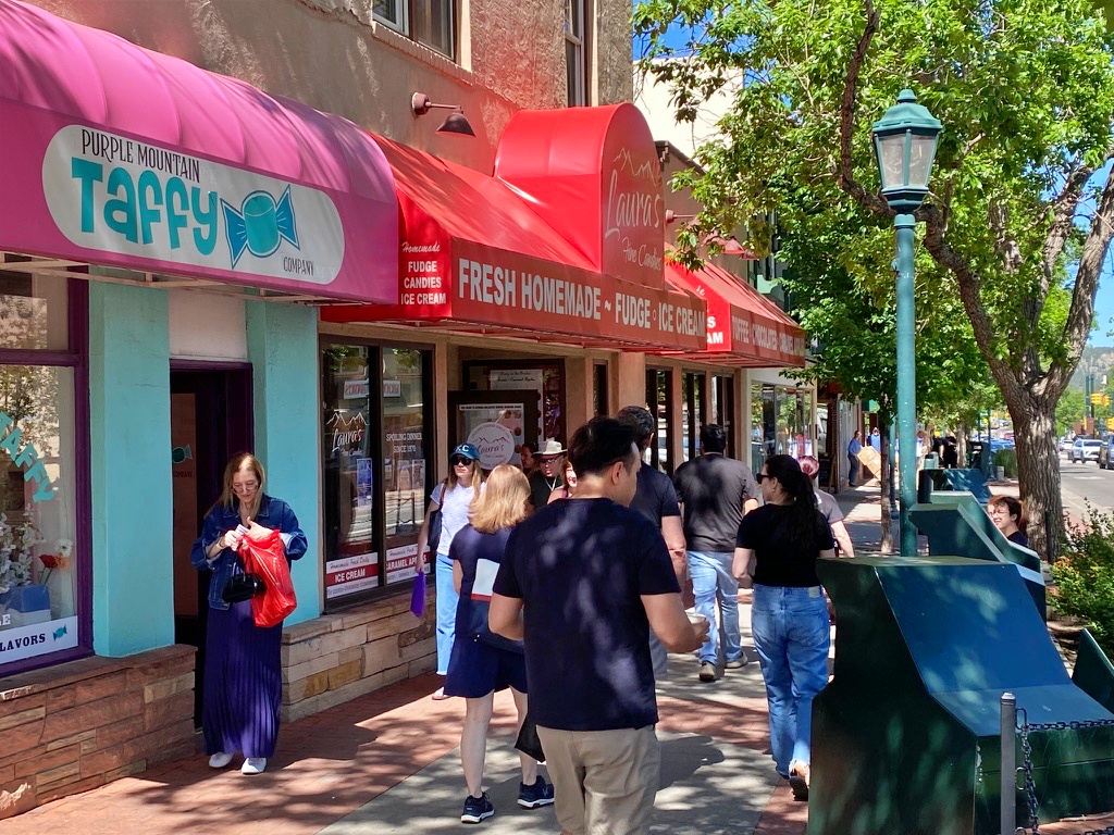 A busy downtown street lined with candy and fudge shop storefronts with colorful awnings, pedestrians walking along the sidewalk, and trees and mountains in the background