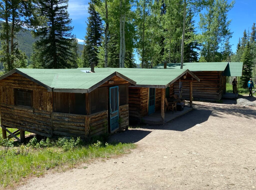 A row of connected rustic log cabins with green roofs and screened porches, one labeled "Rose," set among aspen and pine trees with visitors walking nearby