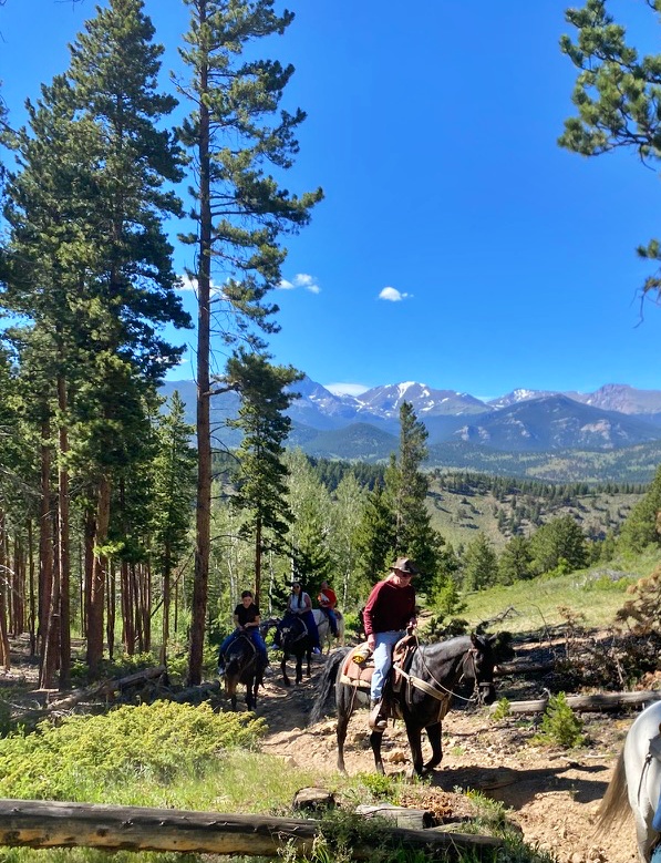 A group of horseback riders on a forested mountain trail, led by a man in a red jacket and cowboy hat, with snow-capped Longs Peak visible in the distance under a blue sky