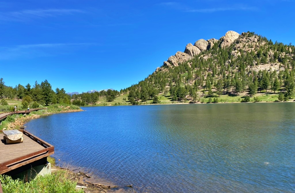 A calm blue lake with a wooden boardwalk and viewing deck on the left, bordered by a rocky, pine-covered ridge and distant mountains under a clear blue sky