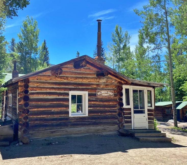 A rustic log cabin with a "Holzwarth Ranch" sign, a small window, a white door, and a metal chimney pipe, surrounded by tall aspen and pine trees under a clear blue sky.