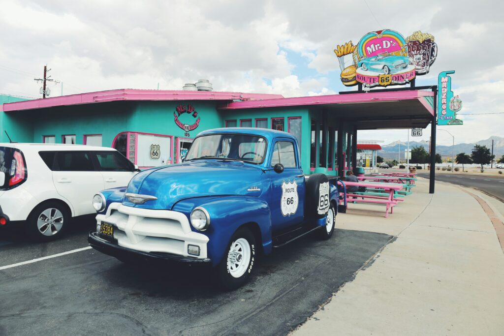 A vintage blue and white 1954 Chevrolet pickup truck with a "Route 66" shield decal parked in front of Mr. D'z Route 66 Diner, a turquoise and pink retro diner in Kingman, Arizona, with a large colorful sign featuring a car, burger, and root beer mug.