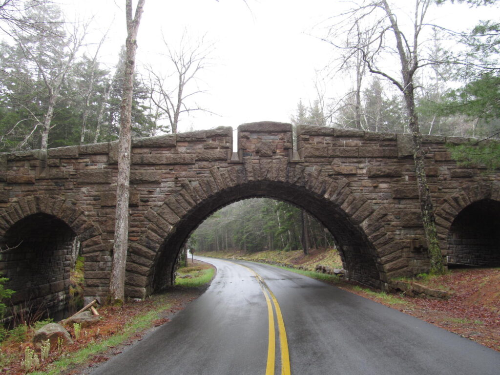 A stone-faced carriage road bridge arching over Park Loop Road in Acadia National Park, surrounded by trees