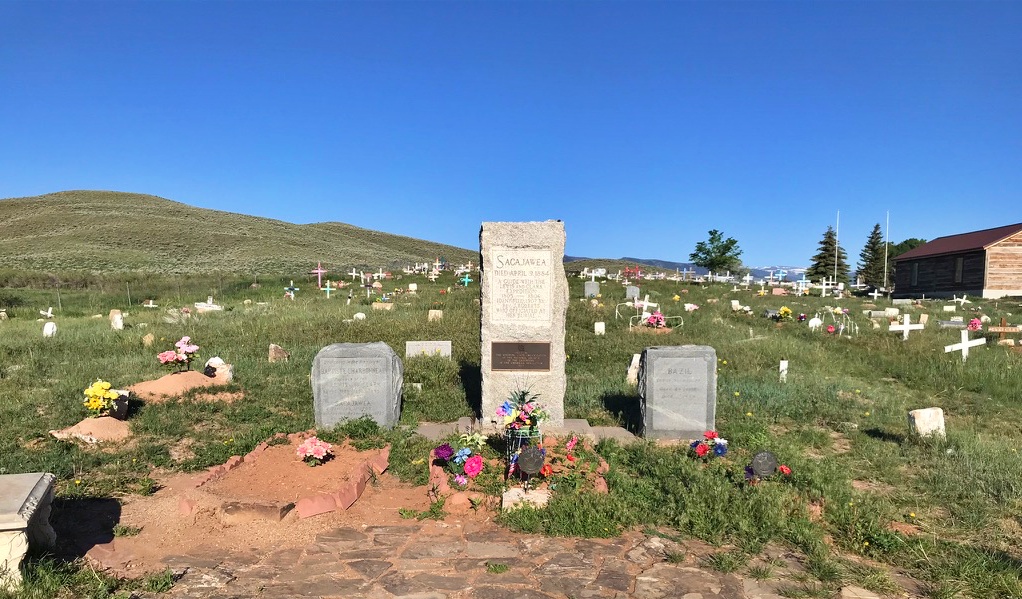 The grave and granite headstone of Sacagawea at the Wind River Reservation cemetery in Fort Washakie, Wyoming. The tall stone monument is surrounded by colorful flowers and flanked by smaller headstones, including those of Toussaint Charbonneau Jr. and Bazil, set against a wide open Wyoming landscape with rolling hills and a vivid blue sky. The Eastern Shoshone oral tradition holds that Sacagawea lived to nearly 100 and is buried here, far from where most historians believe she died