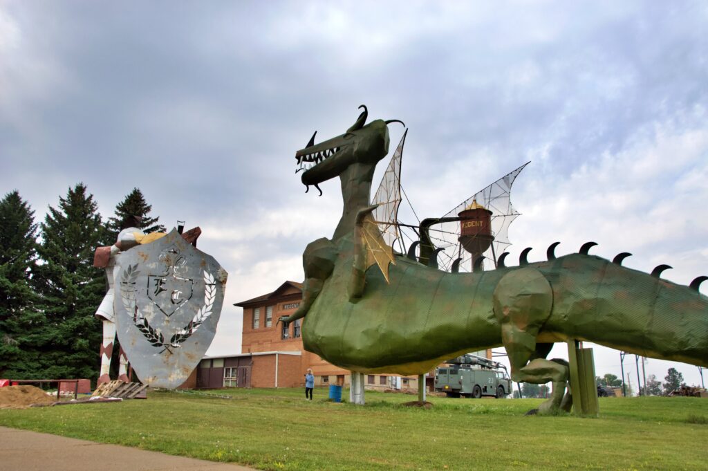 Two giant metal sculptures facing each other across a long sword — a knight in armor on the left and a fanged dragon on the right — towering over a brick schoolhouse, water tower, and parked vehicles for scale, under a cloudy sky