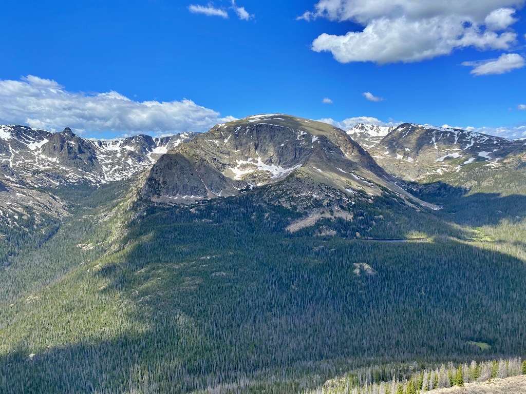 A rugged, snow-streaked mountain peak rising above a dense forested valley, seen from a high overlook under a blue sky with scattered clouds