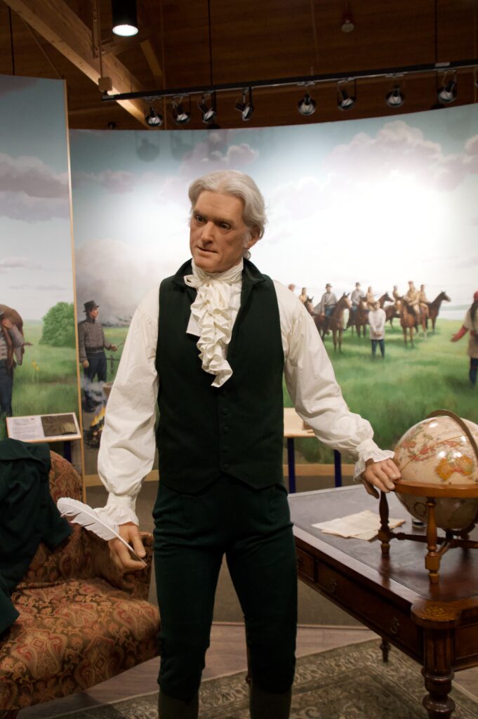 A lifelike animatronic figure of President Thomas Jefferson at the Lewis & Clark Interpretive Center in Sioux City, Iowa. The figure holds a quill pen and stands beside a globe and writing desk, with a panoramic mural depicting the Corps of Discovery on the Great Plains in the background — representing Jefferson's vision that launched the expedition in 1804