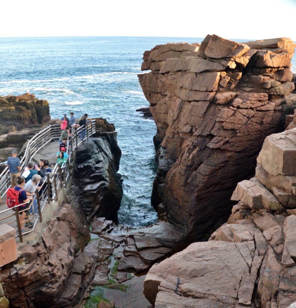 Tourists standing along a railed viewing platform above Thunder Hole, a narrow rock chasm on the Acadia National Park coastline