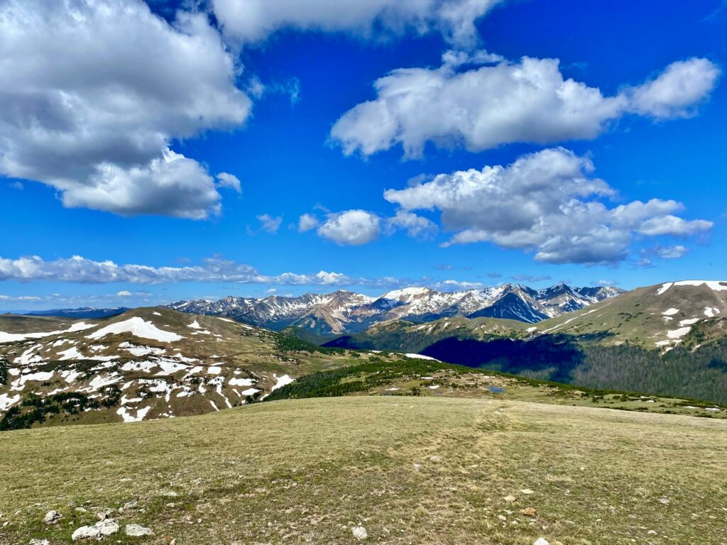 A wide alpine tundra landscape with short green grass in the foreground, rolling snow-patched hills in the middle distance, and the jagged, snow-capped peaks of the Gore Range under a blue sky with scattered white clouds