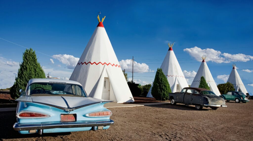 A row of concrete tepee-shaped motel rooms at the Wigwam Motel in Holbrook, Arizona, with a vintage light blue car parked in the foreground