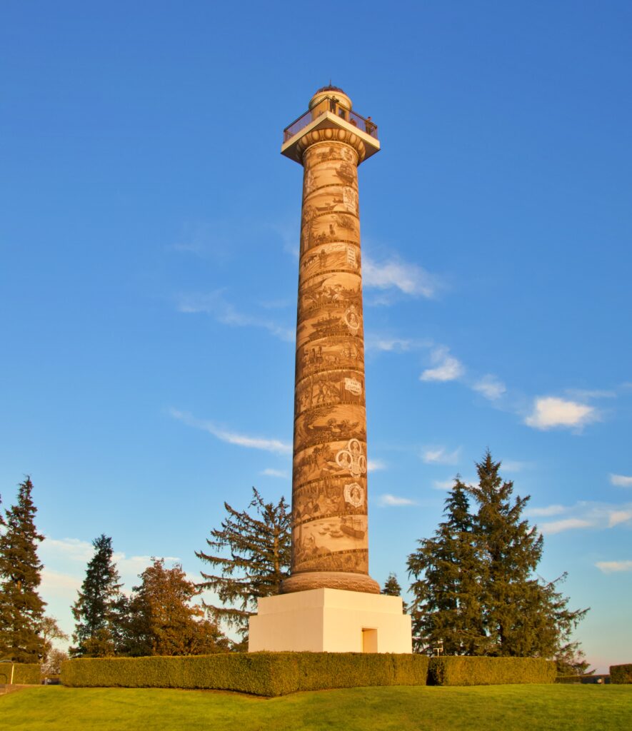 The Astoria Column rises 125 feet above Astoria, Oregon, against a vivid blue sky bathed in warm golden light. The tall concrete column is wrapped in a continuous spiral frieze depicting scenes from Pacific Northwest history including the Lewis and Clark Expedition's arrival at the Pacific coast in 1805. Evergreen trees and a manicured hedge surround the base, and visitors can be seen on the observation deck at the top