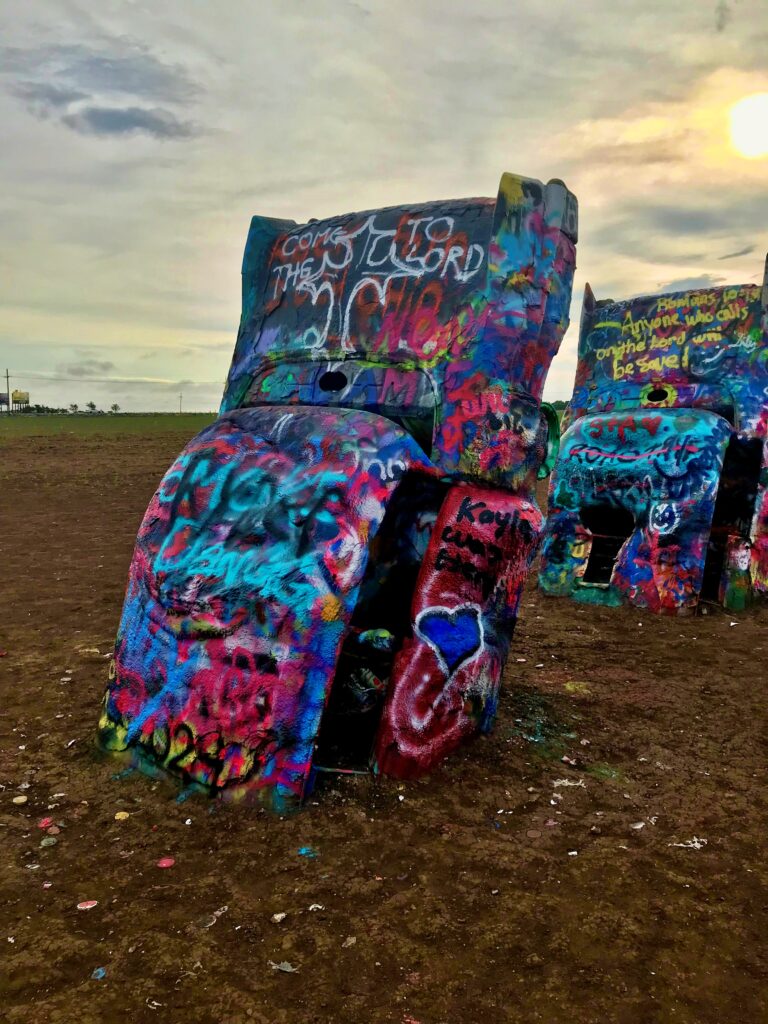 A spray-painted Cadillac half-buried nose-down in a dirt field at Cadillac Ranch, covered in colorful graffiti with more painted cars visible in the background