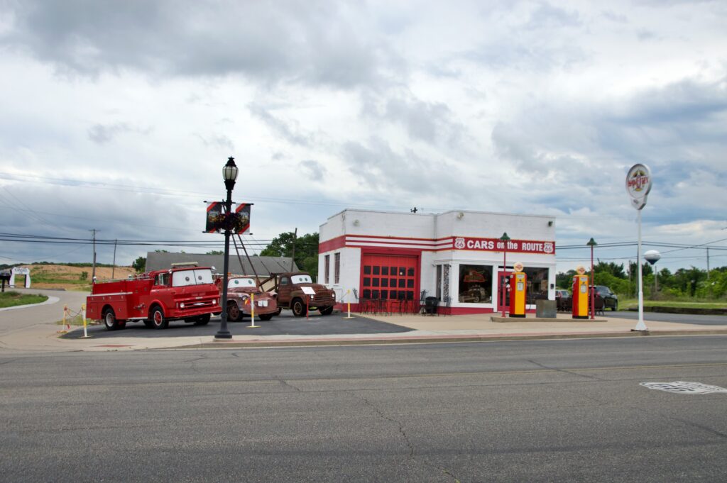 A restored red and white Route 66 gas station called "Cars on the Route" in Galena, Kansas, with a vintage fire truck and two rusted vehicles styled after characters from the movie Cars parked out front