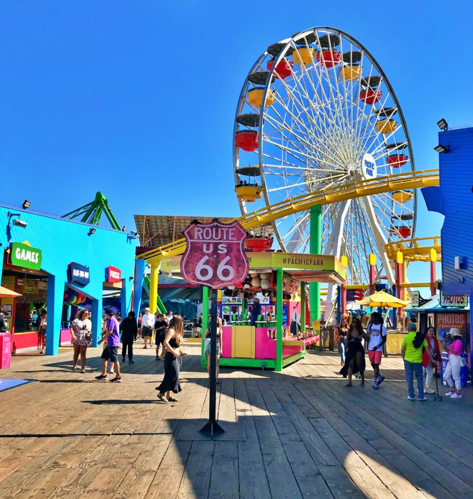 A weathered pink Route 66 shield sign stands on the wooden planks of the Santa Monica Pier, with a large colorful Ferris wheel, bright blue amusement park buildings, and visitors walking under a clear blue sky