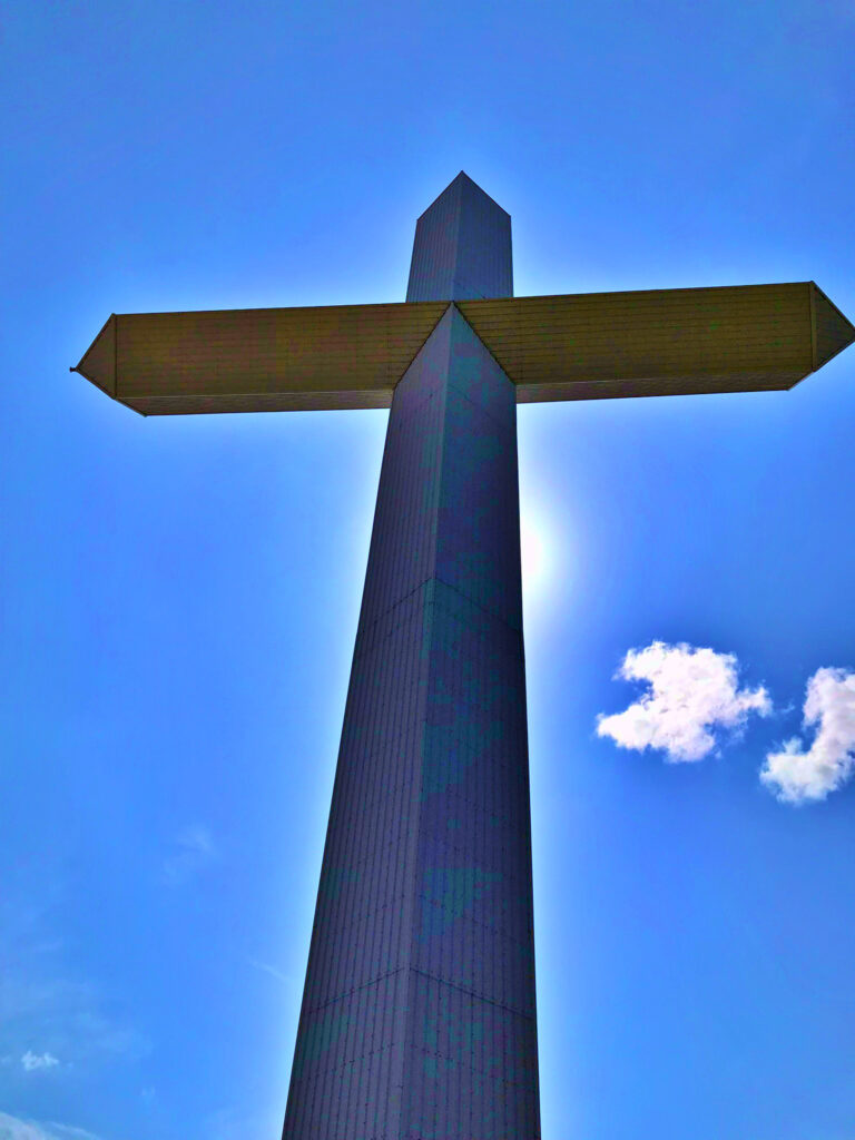 A massive stainless steel cross towering against a bright blue sky with scattered clouds, photographed from below near Groom, Texas