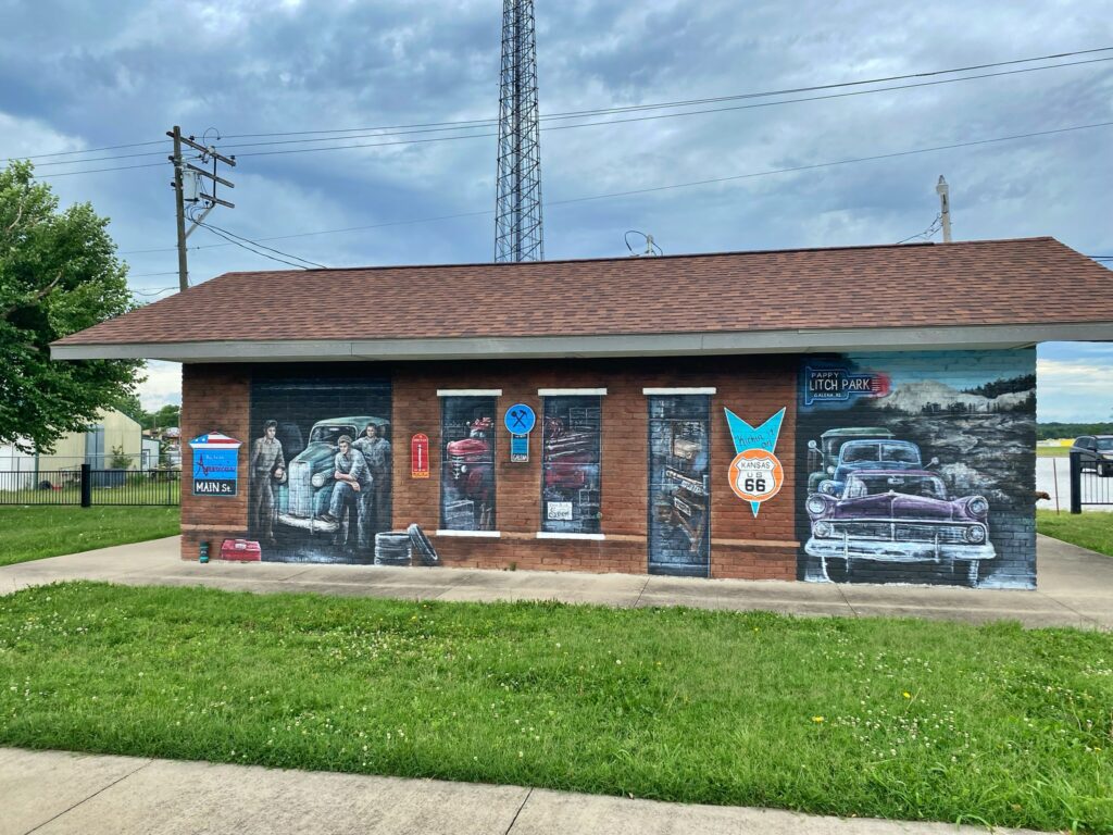A mural on a brick building at Howard "Pappy" Litch Park in Galena, Kansas, depicting vintage mechanics, classic cars, and a Route 66 shield