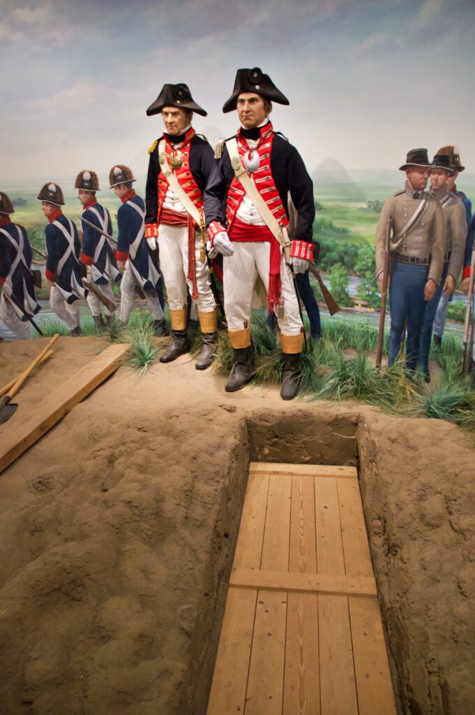 A museum diorama depicting the burial of Sergeant Charles Floyd at the Lewis & Clark Interpretive Center in Sioux City, Iowa. Two lifelike figures of Lewis and Clark in full military dress uniform stand solemnly beside an open grave with a wooden coffin, set against a panoramic mural of the Great Plains landscape. Floyd was the only member of the Corps of Discovery to die during the 8,000-mile expedition, succumbing to what is believed to have been a ruptured appendix on August 20, 1804