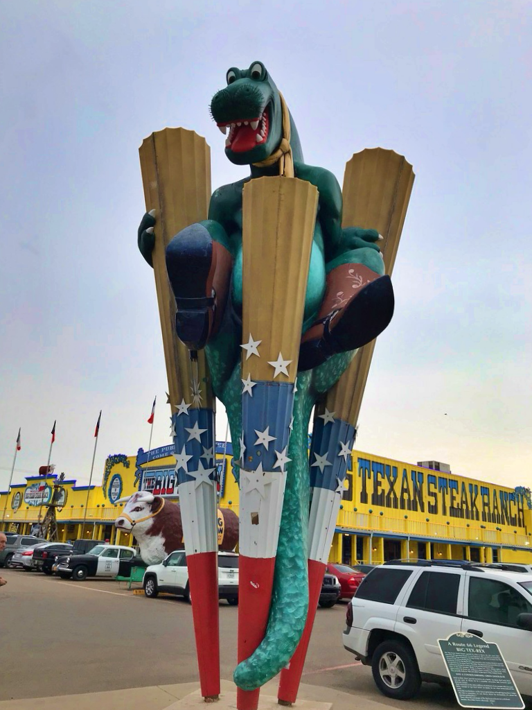 A large green dinosaur statue wearing cowboy boots outside the Big Texan Steak Ranch in Amarillo, Texas, with the restaurant's yellow building in the background