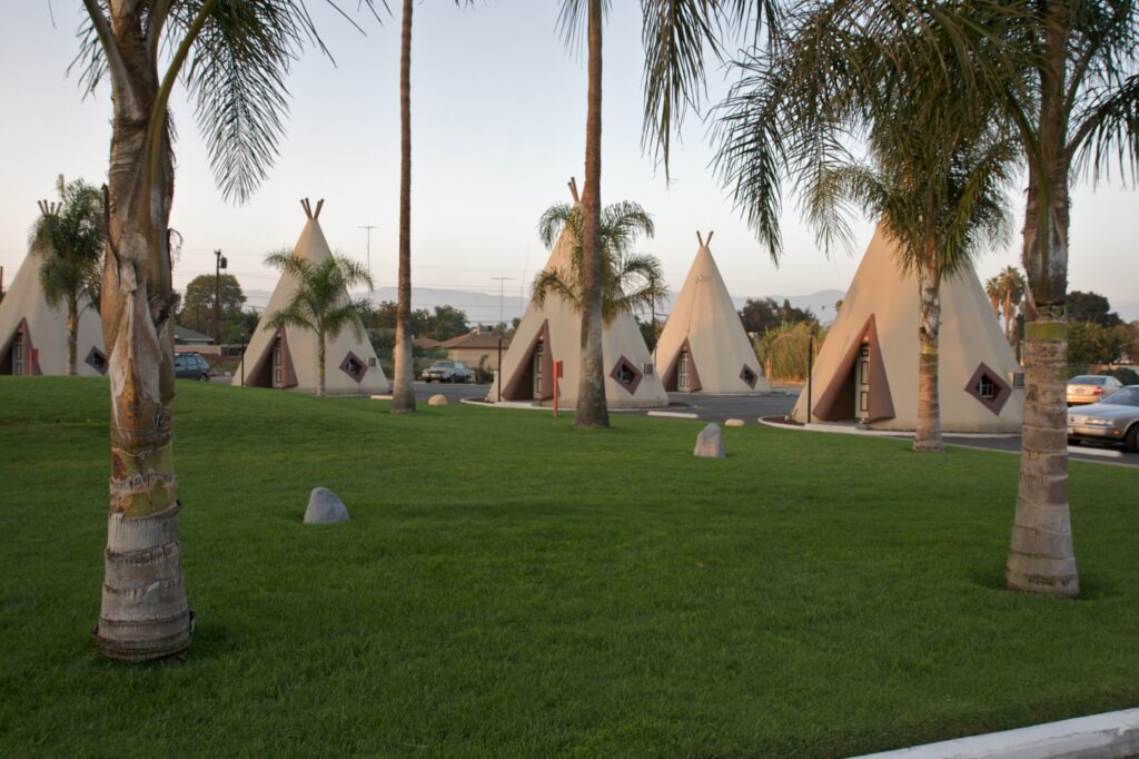 Several large, cream-colored concrete teepee cabins stand on a green lawn interspersed with tall palm trees under a soft, hazy sky