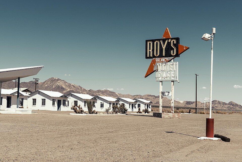 The vintage 'Roy's Motel Cafe' neon sign towering over a row of white motel cabins in Amboy, California, with desert mountains in the background