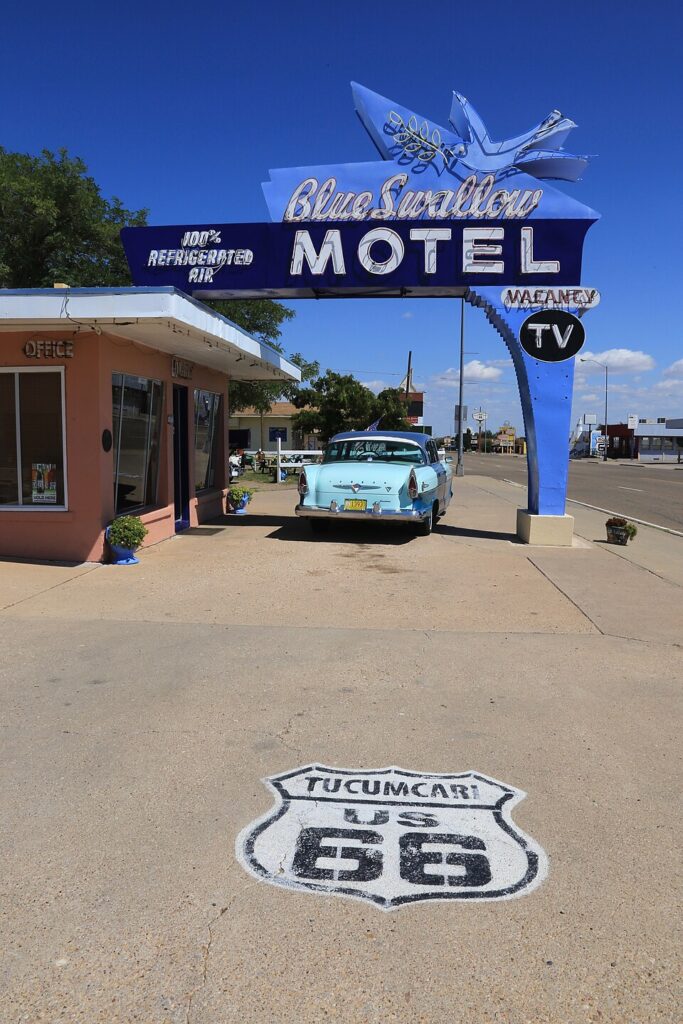 The neon Blue Swallow Motel sign arching over the entrance in Tucumcari, New Mexico, with a vintage car parked beneath it and a Route 66 shield painted on the pavement