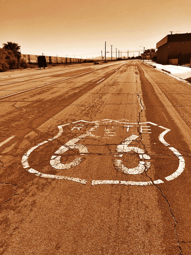 A weathered Route 66 shield painted on cracked desert pavement in Needles, California, stretching toward the horizon under a warm, sepia-toned sky