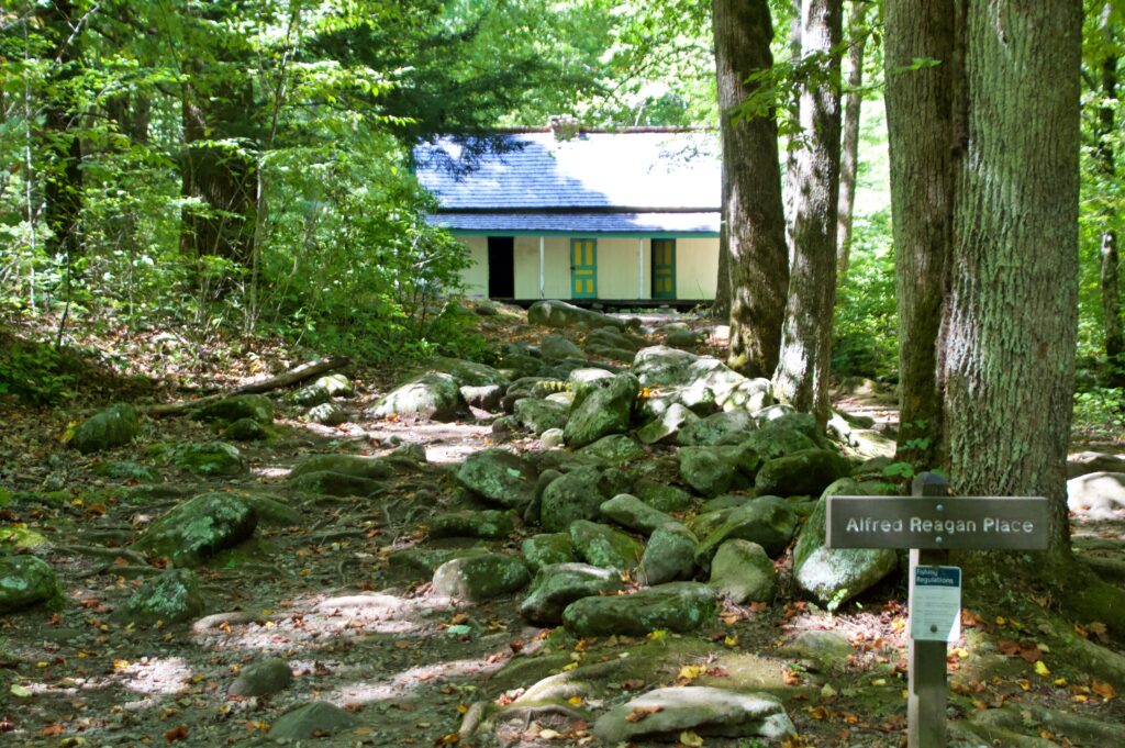 A white painted historic cabin surrounded by trees, viewed from a rocky forest path, with a "Fishing Regulations" posted on a sign that says "Alfred Reagan Place"