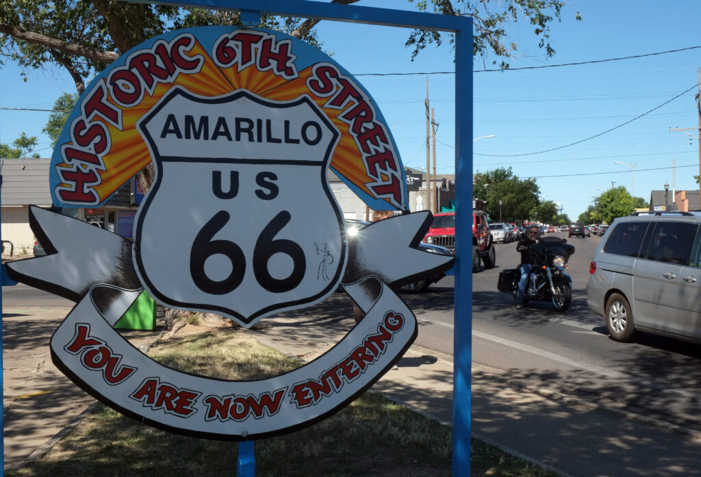 A colorful "Historic 6th Street Amarillo US 66, You Are Now Entering" sign, with a motorcyclist riding past on the street behind it