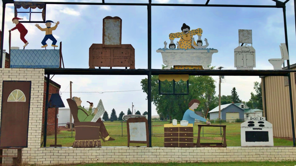 A large flat metal cutaway sculpture of a two-story house showing painted figures in different rooms — children jumping on the roof, a woman bathing two kids in a tub, a man reading a newspaper in a recliner, and a woman cooking at a stove.