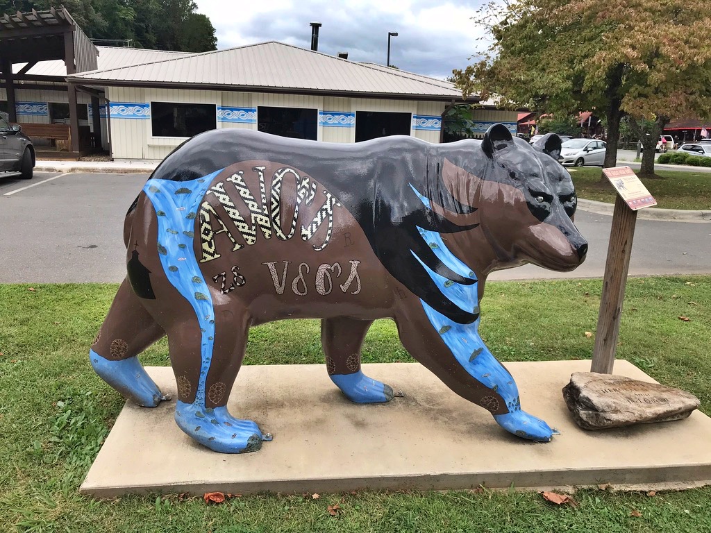 A brown and blue bear statue painted with Cherokee syllabary characters and patterned designs, standing outside a building in downtown Cherokee