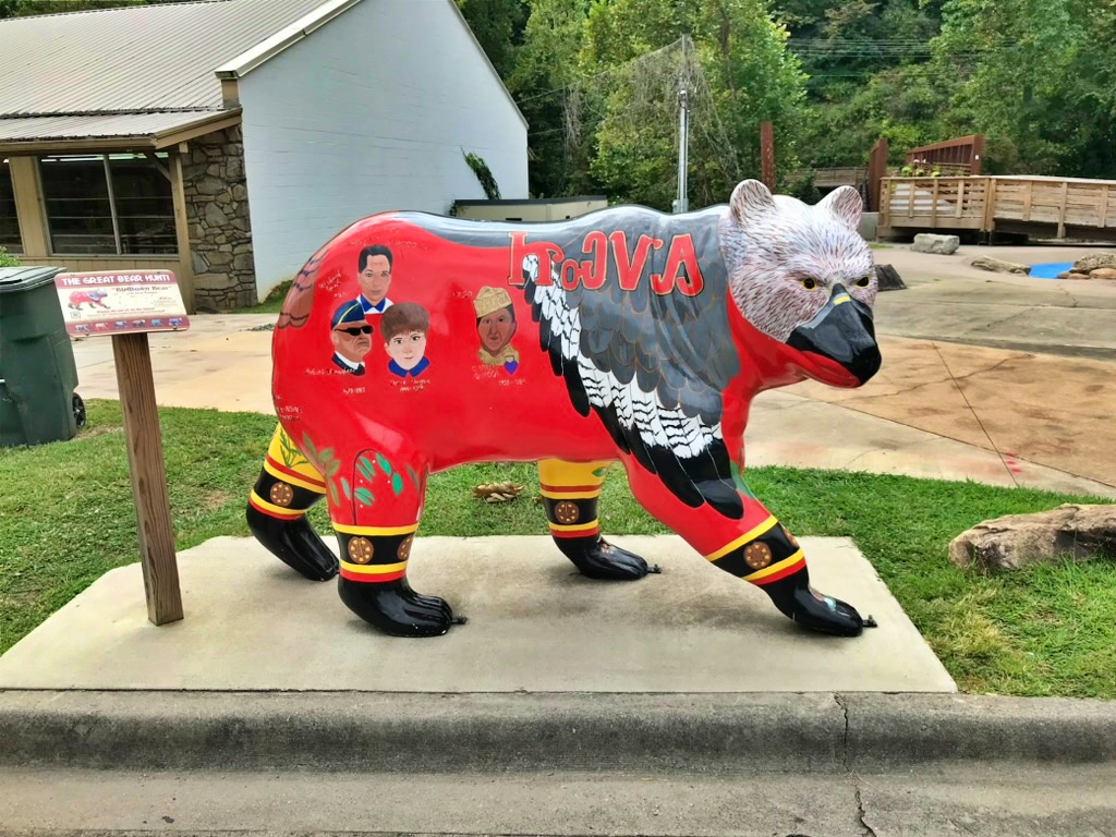 A red bear statue painted with an eagle design and portraits of three people, part of Cherokee's Great Bear Hunt trail