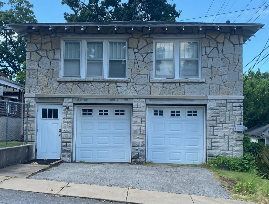A two-story stone and stucco garage apartment with two white garage doors and a small entry door, set on a residential street with trees and power lines in the background
