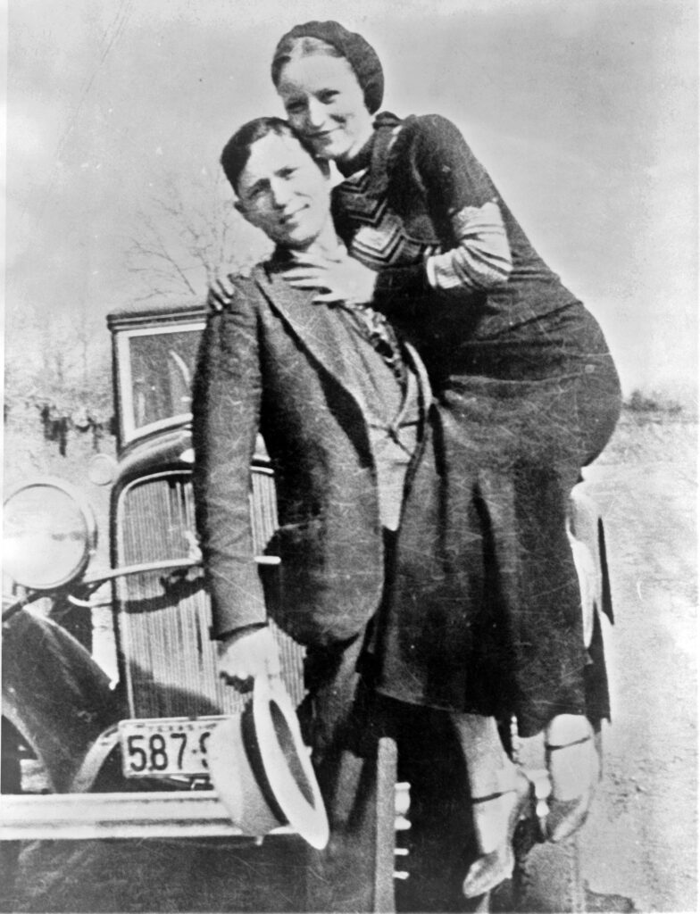 A black-and-white photograph of a man and woman posing in front of a vintage car, the woman with her arm around the man's shoulder, both smiling.