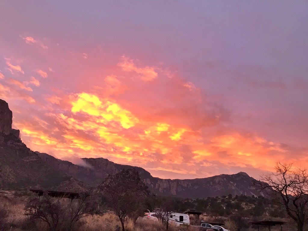 A dramatic orange and pink sunset lights up the sky above Chisos Basin Campground, with an RV and picnic shelters visible in the foreground, at Big Bend National Park