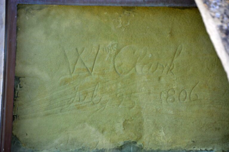 A close-up of William Clark's signature carved into the sandstone face of Pompeys Pillar National Monument near Billings, Montana, reading "W. Clark July 25 1806," photographed through the protective glass case that preserves it. This carving is the only verified original physical evidence of the Lewis and Clark Expedition remaining in place anywhere along the entire 4,900-mile trail, made during the return journey on July 25, 1806.