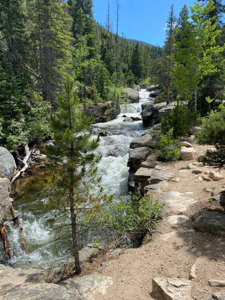 A multi-tiered waterfall cascading over large boulders through a pine forest, with a dirt trail running alongside the creek in the foreground under a clear blue sky