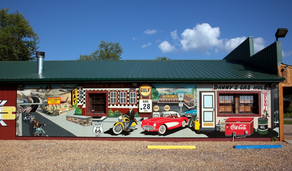 A colorful mural in Cuba, Missouri, depicting a vintage roadside scene with motorcycles, a red Corvette, Carolyn's Cafe, and a Gulf gas station, painted along the length of a building