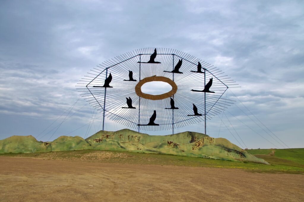 Geese in Flight, a massive scrap metal sculpture of Canada geese in formation surrounding a golden ring, rising above the North Dakota prairie along the Enchanted Highway near Regent. Green painted hills and a cloudy sky frame the artwork, which stands over 110 feet wide and is one of the largest scrap metal sculptures in the world.