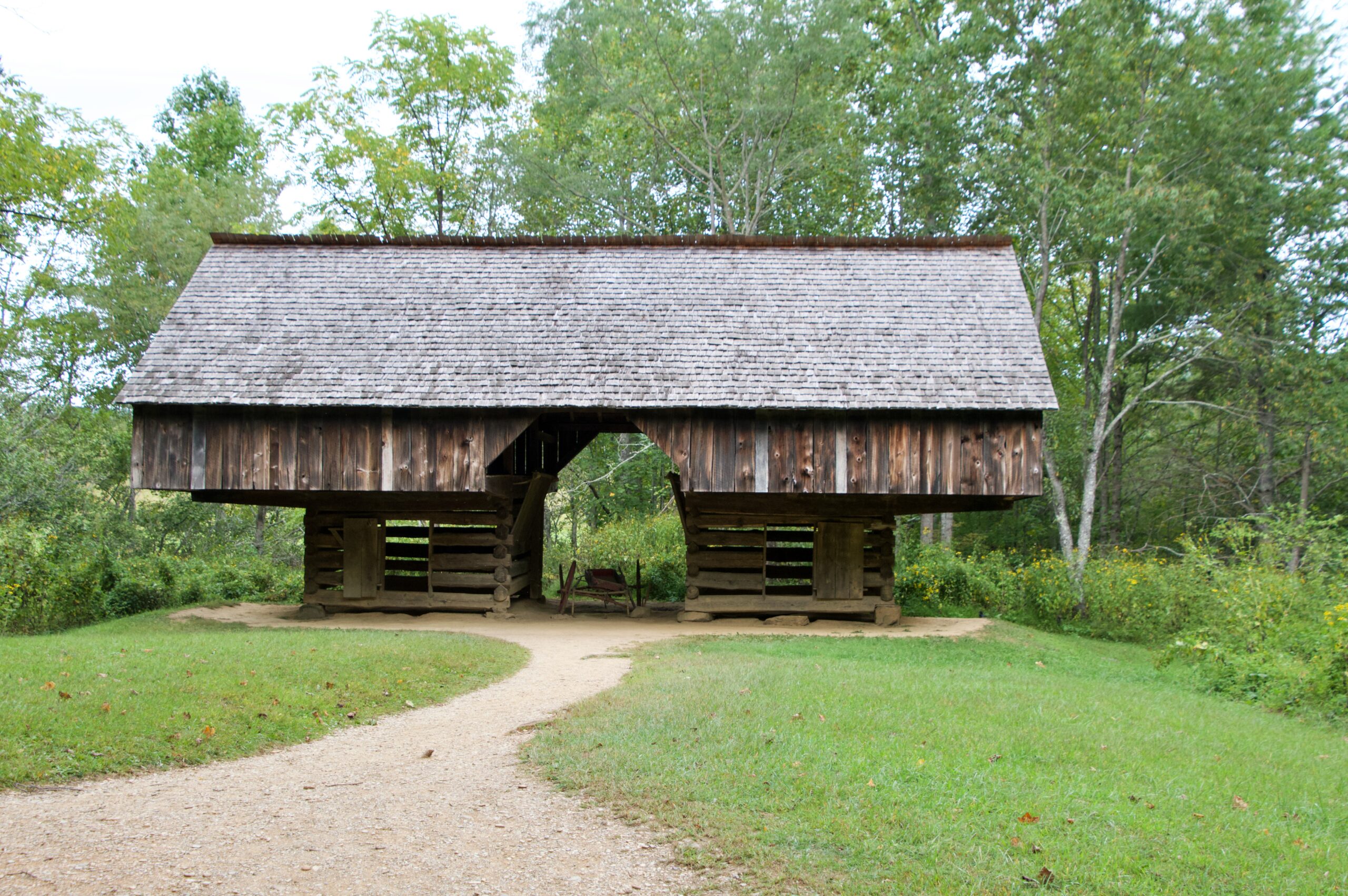 A historic double cantilever barn, a distinctive Appalachian design, at Cades Cove in Great Smoky Mountains National Park, Tennessee. The weathered wooden structure features two log cribs supporting a wide overhanging loft, connected by a central breezeway, surrounded by green meadow and forest