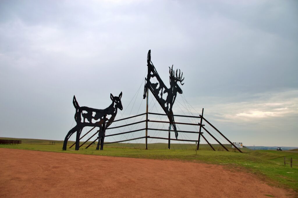 Two large black silhouette metal sculptures of deer, a leaping buck with antlers clearing a wooden fence and a standing doe beside it, set against a cloudy sky and open prairie