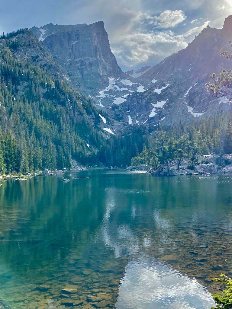 A turquoise alpine lake surrounded by pine trees and rocky shoreline, with the steep, snow-streaked face of Hallett Peak rising against a partly cloudy sky in the background