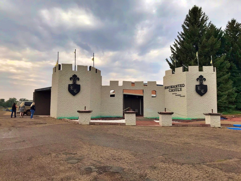 A white castle-style building with turrets, black crest emblems reading "EC," and a sign that says "Enchanted Castle Hotel Tavern Dining," under a cloudy sky